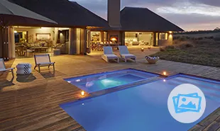 Blue pool with wooden deck and villa in background with lights on